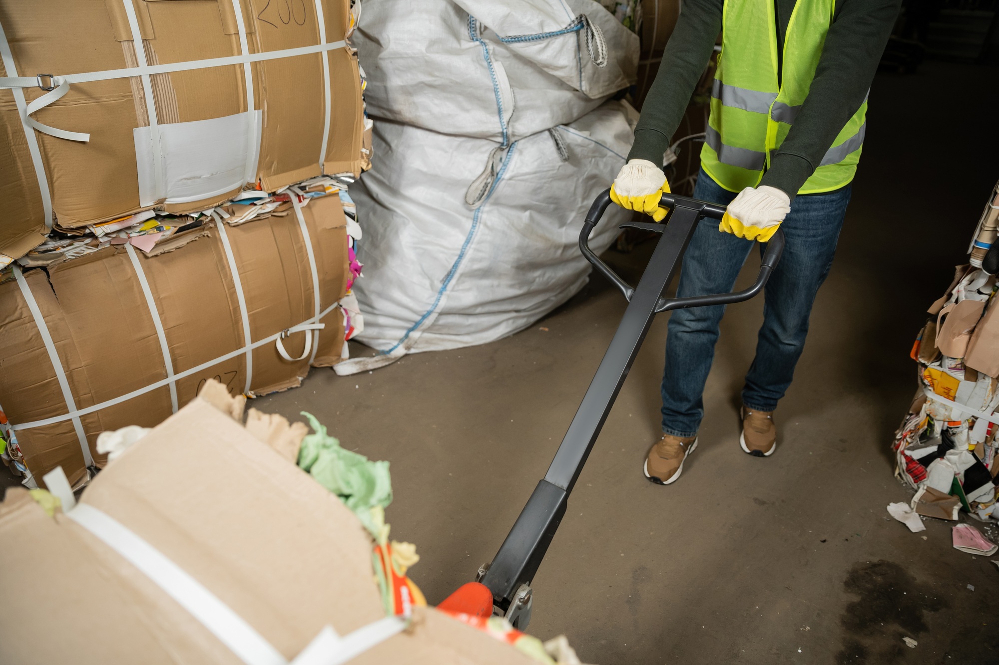 Cropped view of sorter in high visibility vest and gloves using hand pallet truck and moving waste